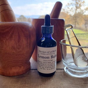 Puede incluir: Una botella de vidrio azul con una etiqueta blanca que dice "Garden of Peper Herbal Apothecary Lemon Balm Tincture". La botella est&aacute; sentada sobre una superficie de madera junto a dos morteros y mazos de madera.