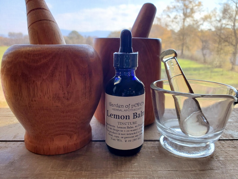 Puede incluir: Una botella de vidrio azul con una etiqueta blanca que dice "Garden of Peper Herbal Apothecary Lemon Balm Tincture". La botella est&aacute; sentada sobre una superficie de madera junto a dos morteros y mazos de madera.