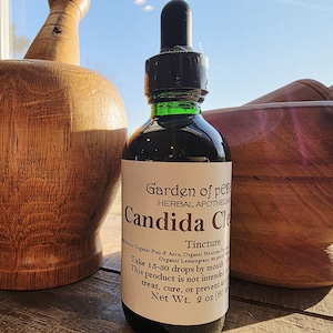 Peut inclure: Une bouteille en verre fonc&eacute; avec un compte-gouttes noir et une &eacute;tiquette blanche indiquant "Candida Cle". La bouteille est &agrave; c&ocirc;t&eacute; de deux outils d'apothicaire en bois. L'&eacute;tiquette indique &eacute;galement "Garden of Pe" et "Herbal Apothecary".