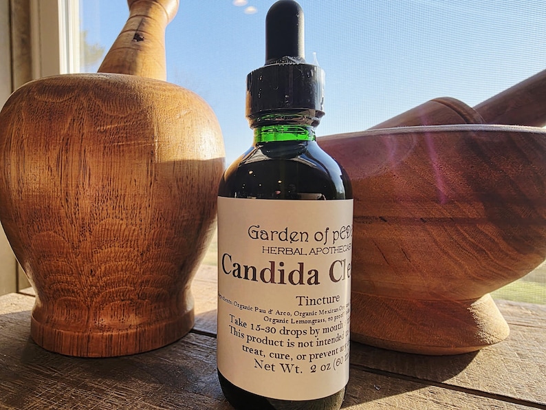 Peut inclure: Une bouteille en verre fonc&eacute; avec un compte-gouttes noir et une &eacute;tiquette blanche indiquant "Candida Cle". La bouteille est &agrave; c&ocirc;t&eacute; de deux outils d'apothicaire en bois. L'&eacute;tiquette indique &eacute;galement "Garden of Pe" et "Herbal Apothecary".