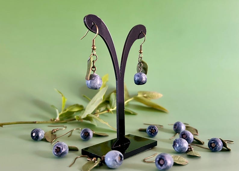 K&ouml;nnte beinhalten: Ein Paar H&auml;ngeohrringe mit einem Blaubeer-Anh&auml;nger und einem gr&uuml;nen Blatt. Die Ohrringe werden auf einem schwarzen St&auml;nder pr&auml;sentiert, um den sich weitere Blaubeer-Anh&auml;nger befinden.