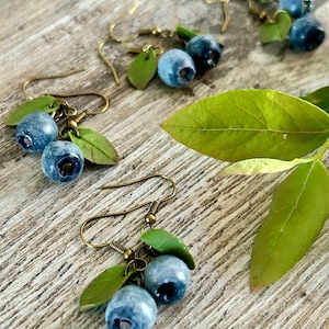 Puede incluir: Un par de pendientes colgantes con dos abalorios azules de arándanos y hojas verdes en cada pendiente. Los pendientes están hechos de metal de color bronce.