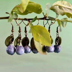 K&ouml;nnte beinhalten: Ein Paar Ohrringe mit einem Blaubeer-Design. Die Ohrringe sind aus Metall und haben ein blaues und gr&uuml;nes Farbschema.