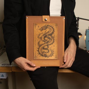 Peut inclure: Un porte-documents en cuir marron avec une illustration détaillée d'un dragon. Le porte-documents a une fermeture à bouton-pression et une petite languette en cuir en bas. Le dragon est dessiné à l'encre noire sur fond beige.