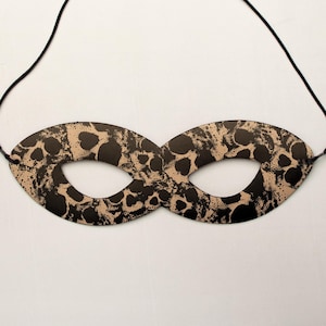 May include: A black and tan masquerade mask with an oval shape and eye openings. The mask has a spotted pattern and is strung with a black cord. The mask is likely made of paper or thin cardboard.