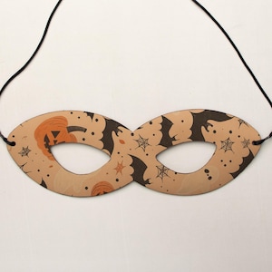 May include: A Halloween-themed eye mask with a tan background. The mask features a repeating pattern of black bats, spiderwebs, and orange pumpkins. A black string is attached to each side of the mask for wearing.