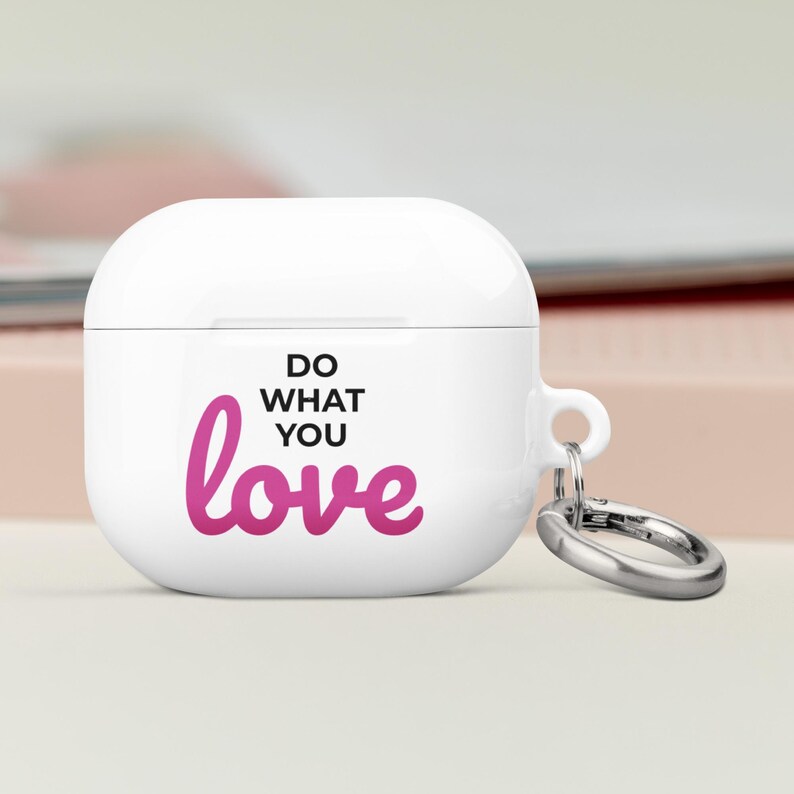 Może przedstawiać: Białe etui na Airpody z r&oacute;żowym napisem "Do what you love" i srebrnym brelokiem.