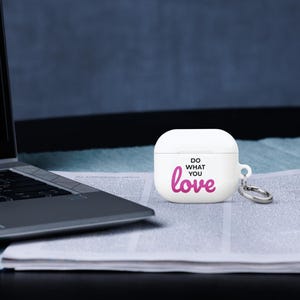 Może przedstawiać: Białe etui na Airpody z r&oacute;żowym i czarnym wzorem z napisem "Do what you love". Etui ma srebrny brelok.
