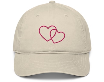 May include: A beige baseball cap with two pink intertwined hearts embroidered on the front.