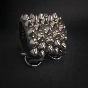 May include: Black leather wristband with rows of silver metal cone studs. The bracelet has two silver metal rings on the bottom. The wristband is a bold accessory.