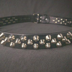 May include: Black leather collar with rows of silver-colored cone studs. The collar has a buckle closure and is designed for wear around the neck. The studs are evenly spaced along the collar's length, creating a bold, edgy look.