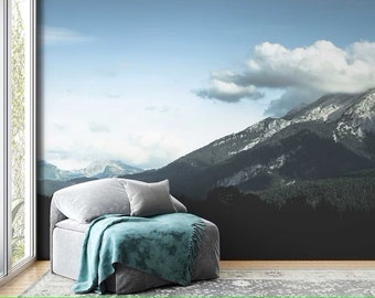 May include: A grey and teal chaise lounge with a throw blanket sits in front of a mountain landscape mural. The mural features a blue sky with white clouds and a mountain range with a forest in the foreground.