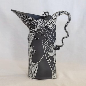 May include: A black ceramic pitcher with a white, stylized profile of a person on the front. The pitcher has a unique handle and is decorated with geometric patterns and floral designs. The spout is pointed and angular.