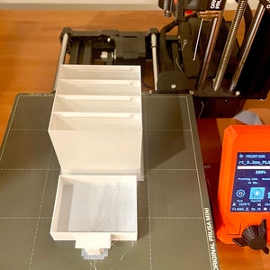 Puede incluir: Un organizador blanco impreso en 3D con cuatro compartimentos se est&aacute; imprimiendo en una impresora 3D Original Prusa Mini negra y gris. La impresora est&aacute; sobre una superficie de madera. La pantalla de la impresora muestra que est&aacute; imprimiendo al 100% y el tiempo de impresi&oacute;n es de 4 h.