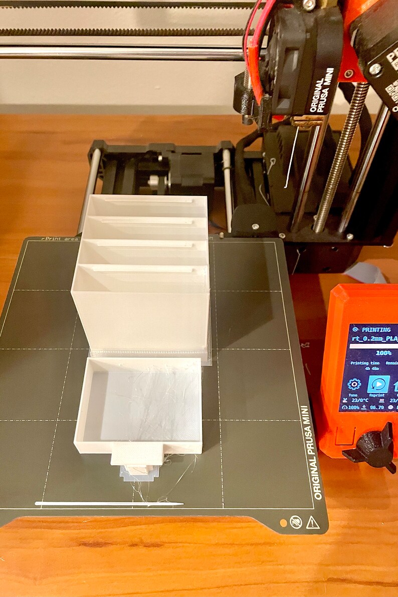 Puede incluir: Un organizador blanco impreso en 3D con cuatro compartimentos se est&aacute; imprimiendo en una impresora 3D Original Prusa Mini negra y gris. La impresora est&aacute; sobre una superficie de madera. La pantalla de la impresora muestra que est&aacute; imprimiendo al 100% y el tiempo de impresi&oacute;n es de 4 h.