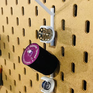 May include: White pegboard organizer with a spool of black polyester thread and a metal bobbin. The thread is labeled "Polyester thread" and is 137 meters long. The organizer is designed for hand and machine sewing.