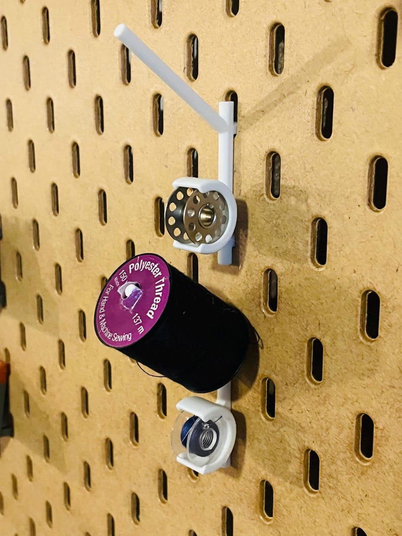 May include: White pegboard organizer with a spool of black polyester thread and a metal bobbin. The thread is labeled "Polyester thread" and is 137 meters long. The organizer is designed for hand and machine sewing.