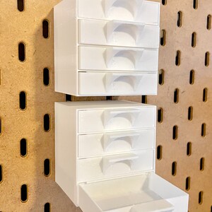 Puede incluir: Organizador de cajones de pl&aacute;stico blanco con seis cajones peque&ntilde;os y un caj&oacute;n m&aacute;s grande. El organizador est&aacute; montado en una pared de tablero de clavijas.