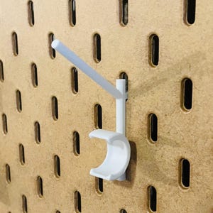 May include: A white pegboard hook with a curved cradle and a straight arm, designed for use with a pegboard. The hook is made of plastic and is mounted on a light brown pegboard with evenly spaced oval holes.