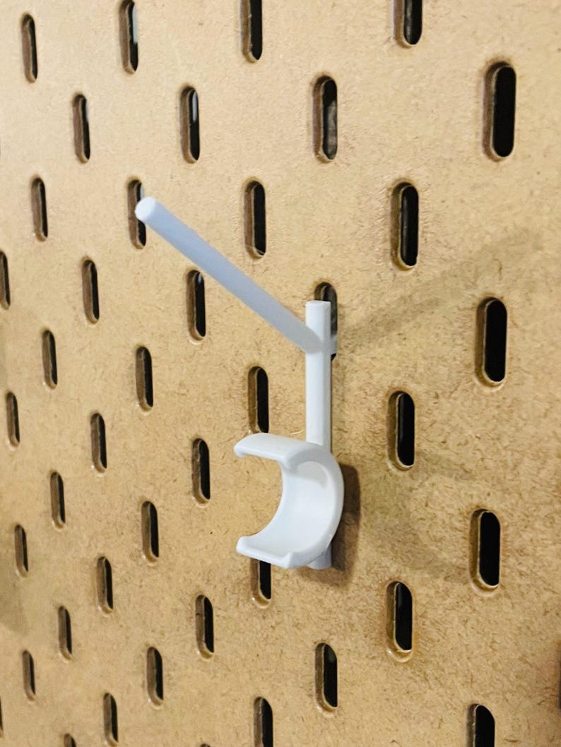 May include: A white pegboard hook with a curved cradle and a straight arm, designed for use with a pegboard. The hook is made of plastic and is mounted on a light brown pegboard with evenly spaced oval holes.
