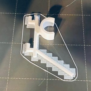 May include: A white 3D-printed object, possibly a custom design, featuring a stylized letter "C" and a staircase-like structure. The object is on a dark surface with grid lines, suggesting a 3D printing bed.