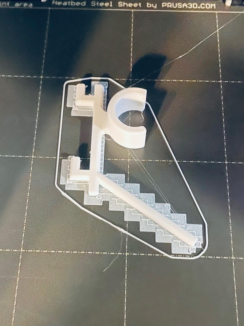 May include: A white 3D-printed object, possibly a custom design, featuring a stylized letter "C" and a staircase-like structure. The object is on a dark surface with grid lines, suggesting a 3D printing bed.