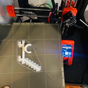 May include: A 3D printer with an orange frame and black components. A gray heatbed steel sheet displays a white 3D-printed model of a staircase and a letter "C". The printer's control panel is visible, displaying text.