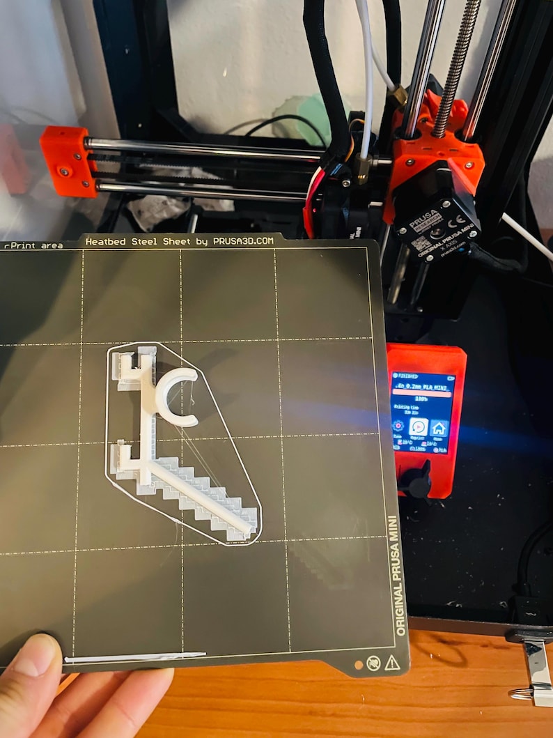 May include: A 3D printer with an orange frame and black components. A gray heatbed steel sheet displays a white 3D-printed model of a staircase and a letter "C". The printer's control panel is visible, displaying text.