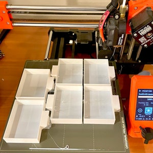 Puede incluir: Una impresora 3D con una cama de impresi&oacute;n blanca y seis peque&ntilde;os objetos rectangulares blancos impresos en ella. La impresora est&aacute; etiquetada como "Original Prusa Mini" y la pantalla muestra "Imprimiendo kadisDrawer 100%".