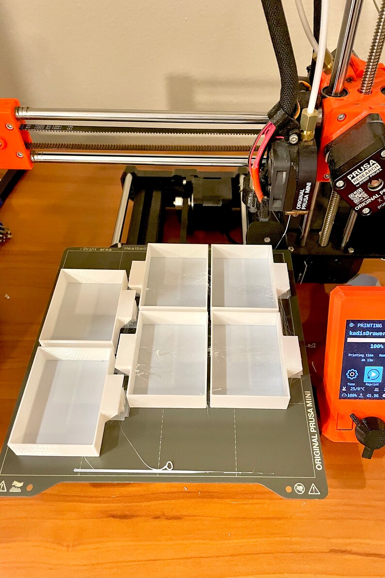 Puede incluir: Una impresora 3D con una cama de impresi&oacute;n blanca y seis peque&ntilde;os objetos rectangulares blancos impresos en ella. La impresora est&aacute; etiquetada como "Original Prusa Mini" y la pantalla muestra "Imprimiendo kadisDrawer 100%".