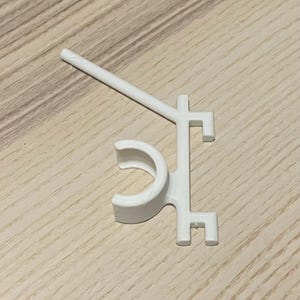 May include: A white plastic clip with a curved section and a straight arm extending from it. The clip has a unique shape with right angles and a small hook at the end. The object is on a light brown wooden surface.