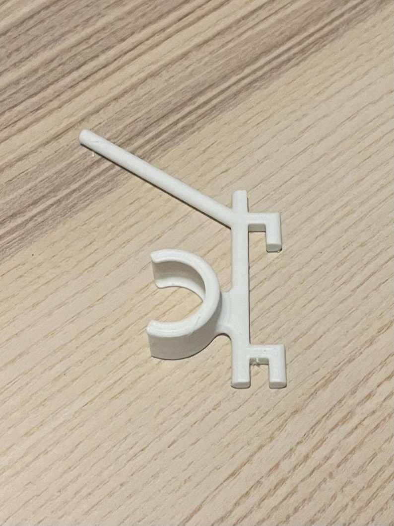 May include: A white plastic clip with a curved section and a straight arm extending from it. The clip has a unique shape with right angles and a small hook at the end. The object is on a light brown wooden surface.