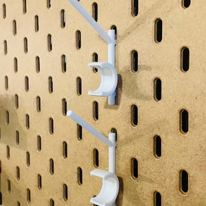 May include: Two white pegboard hooks are mounted on a wooden pegboard. Each hook has a curved cradle and a straight arm extending upwards. The pegboard has evenly spaced, oval-shaped holes.