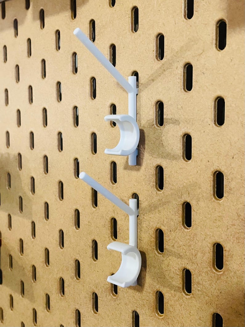 May include: Two white pegboard hooks are mounted on a wooden pegboard. Each hook has a curved cradle and a straight arm extending upwards. The pegboard has evenly spaced, oval-shaped holes.