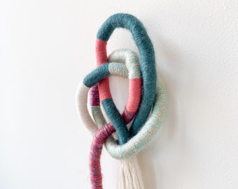 May include: A wall hanging made of woven yarn in teal, pink, and white. The yarn is knotted into a decorative design with a long tassel.