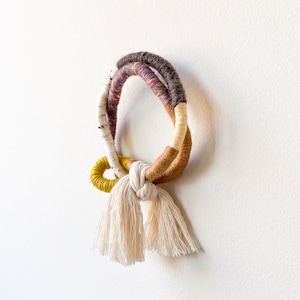 May include: A colourful macrame wall hanging with a circular shape and tassels. The hanging is made with brown, yellow, white and purple yarn.