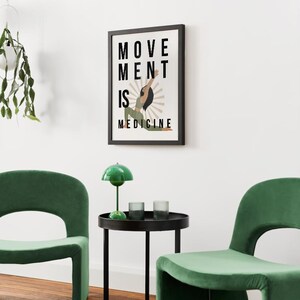 Op de afbeelding: Een ingelijste kunstafdruk met de tekst "MOVEMENT IS MEDICINE". Het kunstwerk toont een persoon in een yogahouding met een sunburst-ontwerp. Twee groene stoelen en een kleine tafel met een groene lamp en glazen staan op de voorgrond.