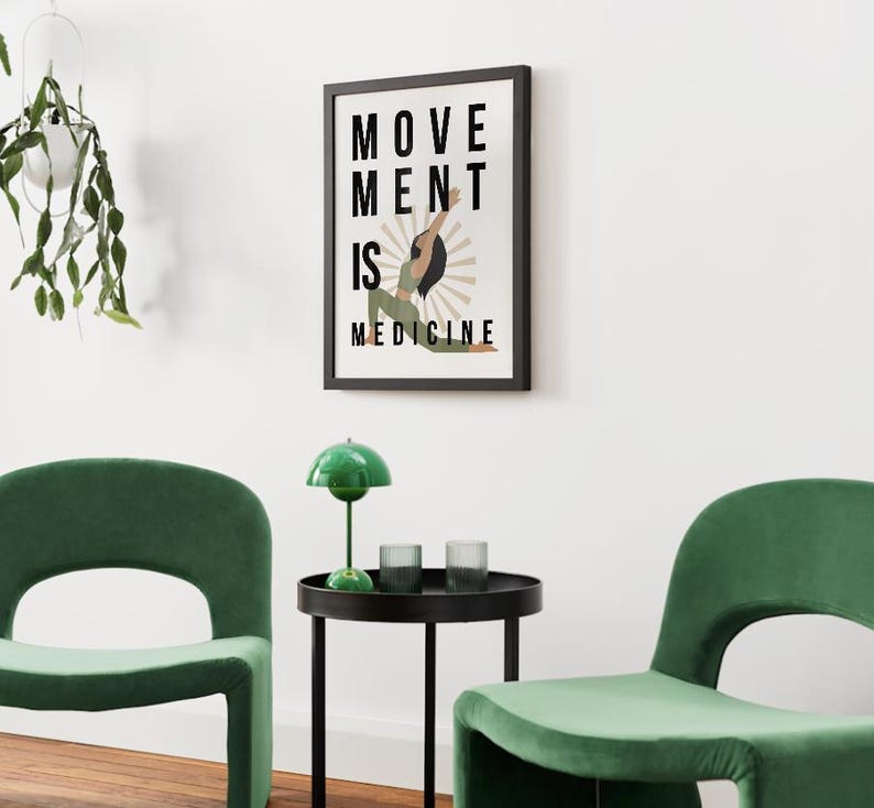 Op de afbeelding: Een ingelijste kunstafdruk met de tekst "MOVEMENT IS MEDICINE". Het kunstwerk toont een persoon in een yogahouding met een sunburst-ontwerp. Twee groene stoelen en een kleine tafel met een groene lamp en glazen staan op de voorgrond.