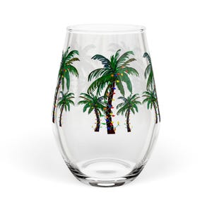 May include: A clear stemless wine glass with a palm tree design. The green palm trees are decorated with colorful Christmas lights. The glass is cylindrical and has a wide base.