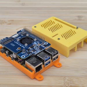 May include: An orange and yellow electronic device with a blue circuit board and silver components. The device has multiple ports and a fan. The text "POE M.2 HAT+" is visible on the blue circuit board. A yellow cover is placed next to the device.