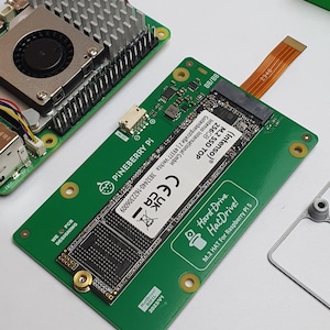 May include: A green Pineberry Pi circuit board with a mounted M.2 SSD, a fan, and a heat sink. The board has the text "Hard Drive. HatDrive! M.2 HAT For Raspberry Pi 5". A ribbon cable connects to the SSD.