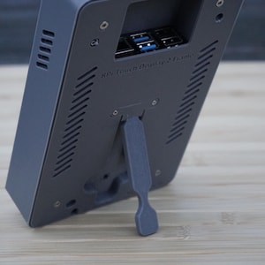 Puede incluir: Un dispositivo rectangular gris con un soporte incorporado, con un área empotrada con puertos USB azules. El dispositivo tiene ranuras de ventilación y el texto "RPi Touch Display 2 Frame".