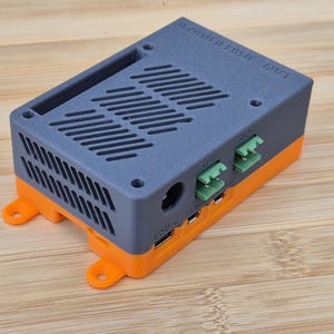 May include: A grey and orange electronic device with vents and ports. The top is grey with horizontal vents, and the bottom is orange. It has several ports and connectors on the sides. The device is sitting on a wooden surface.