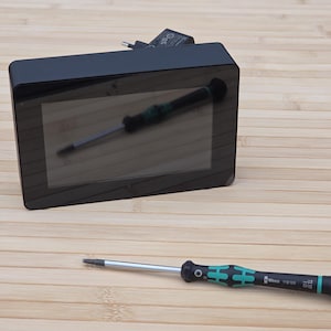 May include: A black electronic device with a reflective screen, and a black and green screwdriver with a silver shaft. The screwdriver is lying on a wooden surface.