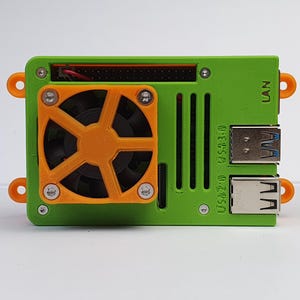 Könnte beinhalten: Ein grünes und orangefarbenes elektronisches Gerät mit einem Lüfter. Das Gerät hat eine rechteckige Form mit orangefarbenen Befestigungsschlaufen. Der Lüfter ist orange und von einem grünen Rahmen umgeben. Es gibt USB-Anschlüsse und das Wort LAN.