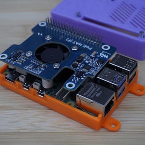 Op de afbeelding: Een blauwe printplaat met een ventilator en de tekst "POE HAT (F)" is gemonteerd op een oranje basis. Een paarse behuizing staat achter de printplaat. De componenten staan op een houten oppervlak.