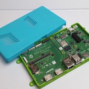 Peut inclure: Une carte Raspberry Pi Compute Module 4 IO, avec un cadre vert et un couvercle bleu turquoise. La carte est verte avec divers composants et ports, dont HDMI et USB. Le couvercle bleu turquoise comporte des fentes d'aération et du texte.