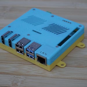 Puede incluir: Un dispositivo electrónico azul y amarillo con múltiples puertos, incluyendo USB y LAN. El dispositivo rectangular tiene una parte superior ventilada y está diseñado para la transferencia de datos y la conectividad. Es probablemente un ordenador pequeño.