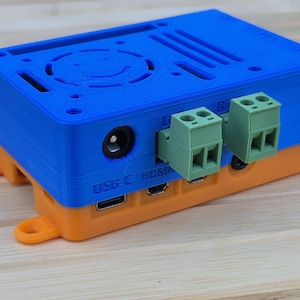 Könnte beinhalten: Ein blau-oranges elektronisches Gerät mit Lüfter und mehreren Anschlüssen. Das Gerät hat grüne Klemmenblöcke und einen USB-C-Anschluss. Das Gerät steht auf einer Holzoberfläche.