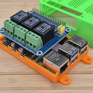 May include: An assembled electronic circuit board with green and black components, including relays and connectors, sits inside an orange plastic case. A green plastic case is in the background. The board has multiple ports and connectors.